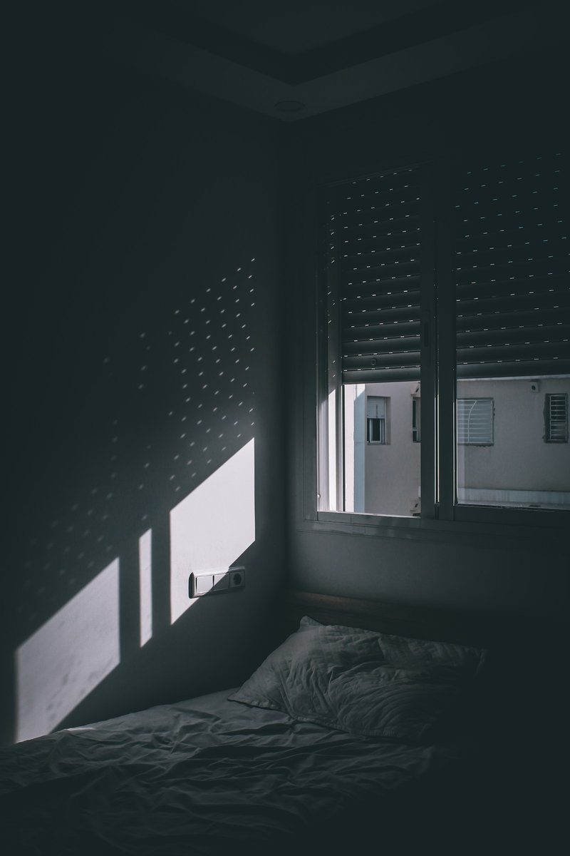 Chambre plongée dans la pénombre, volets roulants presque fermés laissant passer quelques rais de lumière sur un lit défait